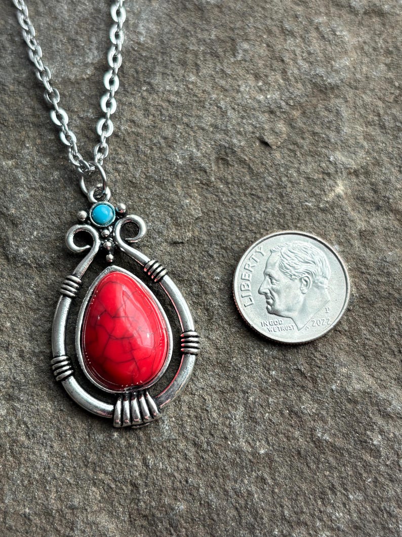 Peut inclure: Un collier en cha&icirc;ne argent avec un pendentif en pierre rouge et blanche. Le pendentif est entour&eacute; d'un cadre argent&eacute; avec une pierre turquoise en haut. Le collier est pr&eacute;sent&eacute; &agrave; c&ocirc;t&eacute; d'une pi&egrave;ce de dix centimes d'euro pour comparaison de taille.