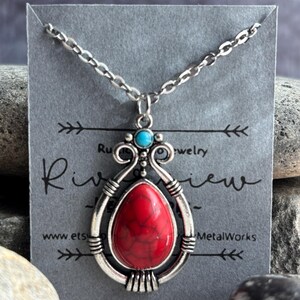 Peut inclure: Un collier en argent avec un pendentif en pierre rouge et blanche. Le pendentif est entour&eacute; d'un motif en filigrane argent&eacute;. Le collier est sur une carte grise avec le texte "Riverview MetalWorks" et une adresse de site web.