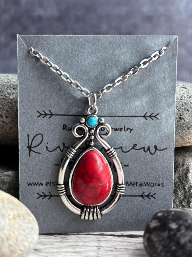 Peut inclure: Un collier en argent avec un pendentif en pierre rouge et blanche. Le pendentif est entour&eacute; d'un motif en filigrane argent&eacute;. Le collier est sur une carte grise avec le texte "Riverview MetalWorks" et une adresse de site web.