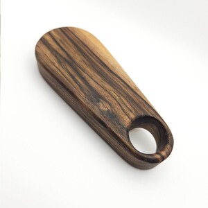 K&ouml;nnte beinhalten: Ein dunkelbrauner Flaschen&ouml;ffner aus Holz mit einem runden Loch im Griff. Das Holz hat ein gestreiftes Muster.