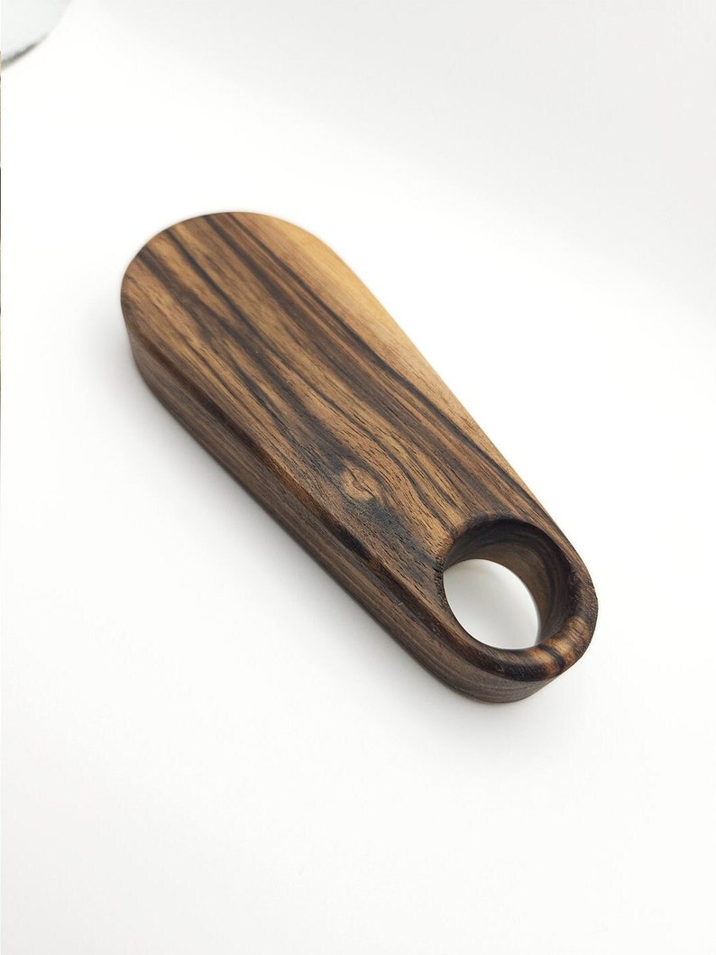 K&ouml;nnte beinhalten: Ein dunkelbrauner Flaschen&ouml;ffner aus Holz mit einem runden Loch im Griff. Das Holz hat ein gestreiftes Muster.