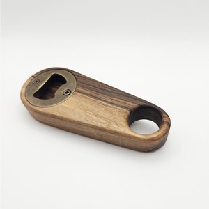 K&ouml;nnte beinhalten: Ein Flaschen&ouml;ffner aus Holz mit einem Messing-Kronkorken&ouml;ffner. Der &Ouml;ffner hat einen Ring am Ende zum einfachen Aufh&auml;ngen.