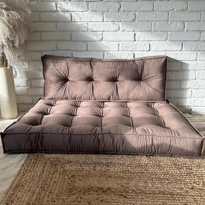 Könnte beinhalten: Ein Bodenkissen-Set in gedämpftem Mauve mit getuftetem Design und mehreren Stoffknöpfen. Das Set besteht aus einem rechteckigen Bodenkissen und einem passenden rechteckigen Rückenkissen, beide mit paspelierten Kanten und einer weichen Textur.
