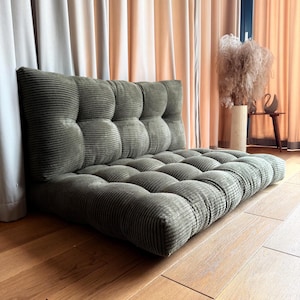May include: An olive green corduroy floor chair with a tufted back and seat. The chair is against a wall and has evenly spaced, rounded tufts. The corduroy fabric has a ribbed texture. The chair's dimensions are in centimetres.