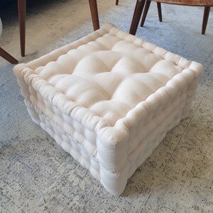 May include: A white, tufted, square floor cushion with a linen-like fabric. The cushion is made of multiple layers and has a thick, comfortable appearance.