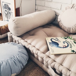 Peut inclure: Un coussin rond bleu clair avec un bord festonné repose sur un coussin de sol en lin beige. Le coussin de sol est surmonté d'un long coussin de soutien en lin beige. Un livre intitulé "Diplodok" avec une illustration de dinosaure bleu est sur le coussin de sol.