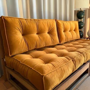 May include: A mustard-colored futon with a tufted design. The futon has a wooden frame and two back cushions. The cushions and seat are made of a soft, velvety material. The futon is in a well-lit room.