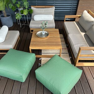 May include: A wooden outdoor furniture set with a sofa, two chairs, a coffee table, and two green ottomans. The furniture is arranged on a wooden deck with a rug in the centre.