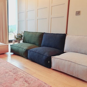 May include: A modular seating arrangement consisting of a light beige ottoman, a dark green floor chair, a dark blue floor chair, and a light beige floor chair. Each piece features a textured fabric finish, square edges, and a low-profile design.