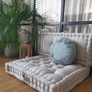 Op de afbeelding: Een groot, lichtgrijs, gestoffeerd vloer kussen met een lichtblauw rond kussen erop. Het kussen ligt op een houten vloer voor een raam met witte gordijnen.
