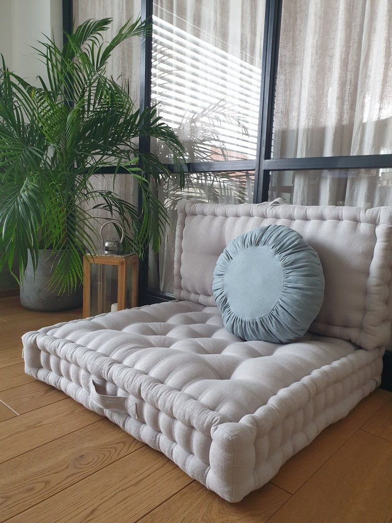 Op de afbeelding: Een groot, lichtgrijs, gestoffeerd vloer kussen met een lichtblauw rond kussen erop. Het kussen ligt op een houten vloer voor een raam met witte gordijnen.