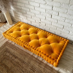 Peut inclure: Un coussin de sol jaune moutarde avec des boutons capitonn&eacute;s et une &eacute;paisse bordure cousue. Le coussin est en velours et repose sur un tapis tiss&eacute;.