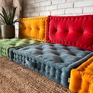 May include: Four floor cushions in different colors, including green, yellow, red, and blue. The cushions are made of a soft, plush fabric and have a tufted design. The cushions are arranged in a row on a woven rug.
