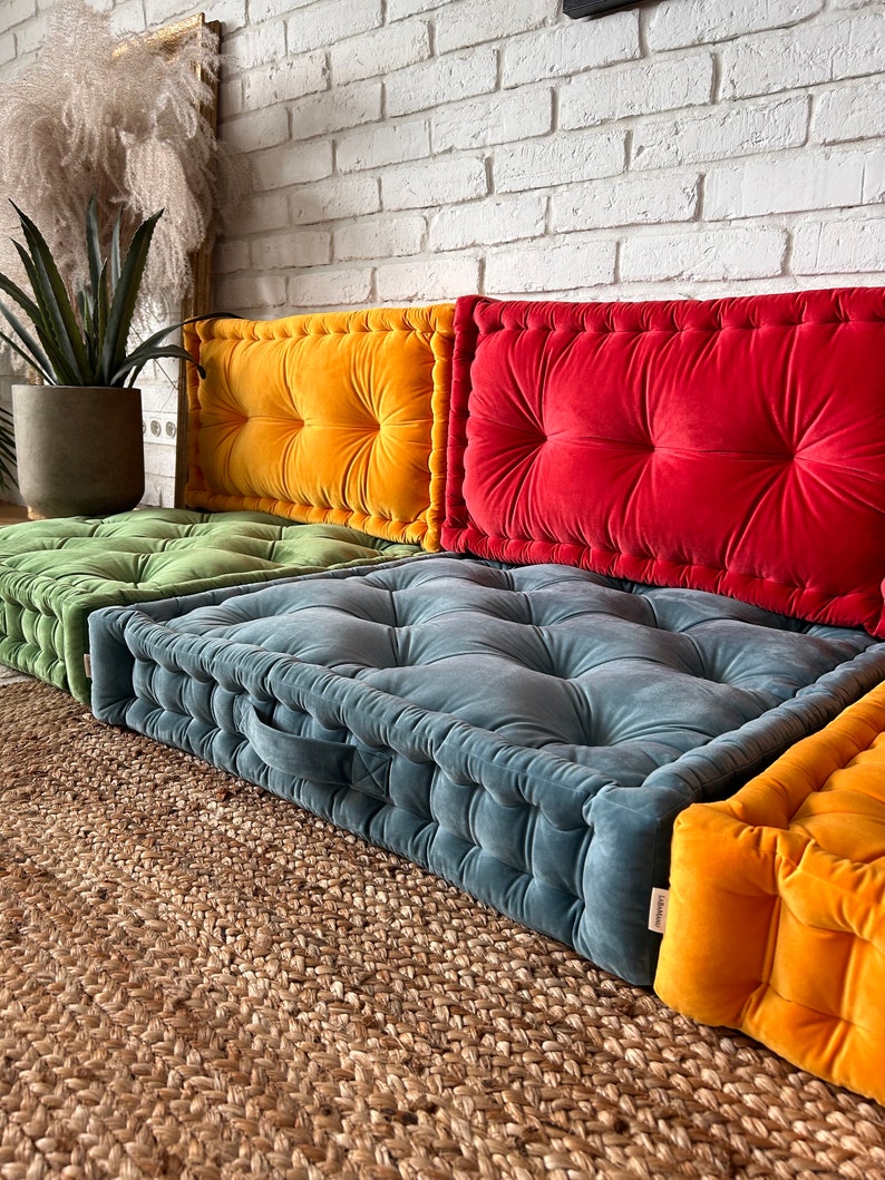 May include: Four floor cushions in different colors, including green, yellow, red, and blue. The cushions are made of a soft, plush fabric and have a tufted design. The cushions are arranged in a row on a woven rug.