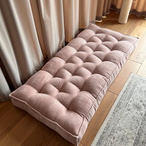 May include: A pink corduroy floor cushion with a tufted design. The cushion is rectangular and has a thick, padded base.