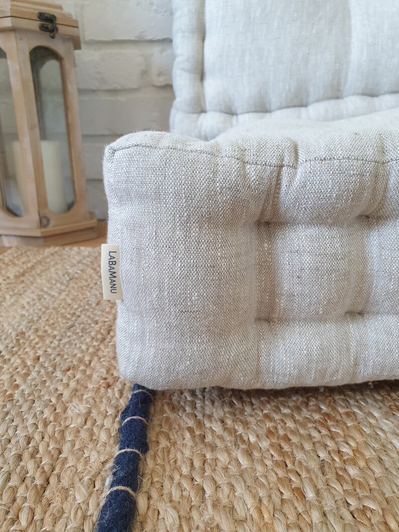 Op de afbeelding: Een close-up van een lichtgrijs linnen vloer kussen met een gestoffeerd ontwerp. Het kussen heeft een label met de tekst "LABAMANU".