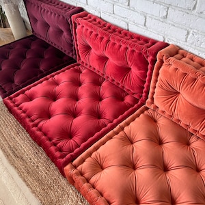 May include: Three floor cushions in shades of burgundy, red, and orange. The cushions are square and have a tufted design with button accents. The cushions are arranged in a row on a woven rug.