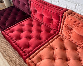 May include: Three floor cushions in shades of burgundy, red, and orange. The cushions are square and have a tufted design with button accents. The cushions are arranged in a row on a woven rug.