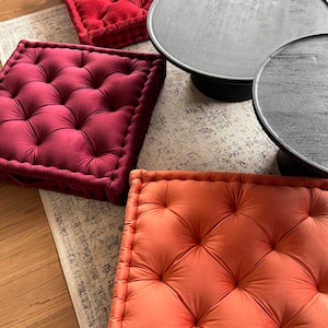 May include: Three square floor cushions with tufted tops. Two cushions are burgundy velvet and one is a burnt orange velvet.