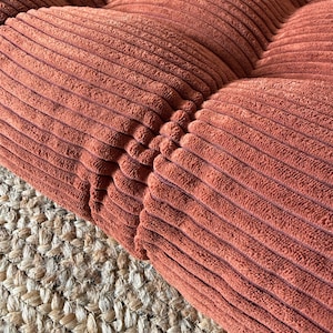 K&ouml;nnte beinhalten: Nahaufnahme eines Abschnitts eines Cord-Kissens in einem warmen, r&ouml;tlich-braunen Farbton. Das Kissen hat ein gestepptes Design mit sichtbaren N&auml;hten. Die Textur des Cordes ist deutlich sichtbar, und das Kissen liegt auf einem gewebten Jute-Teppich.