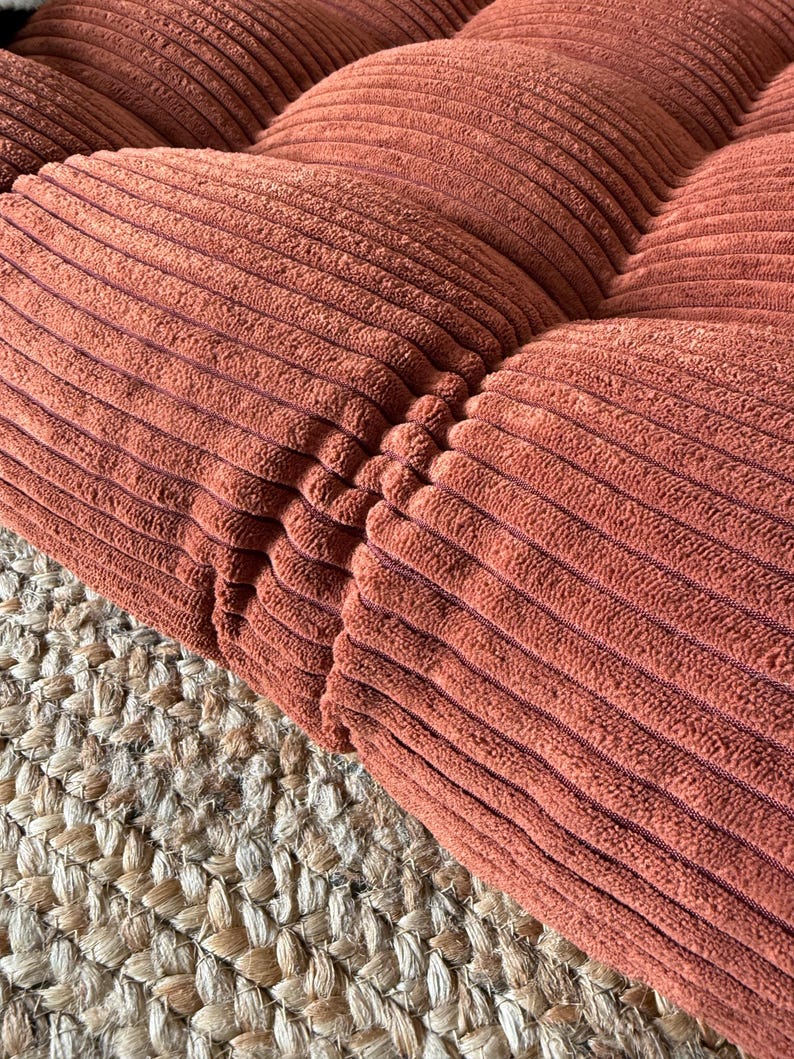 K&ouml;nnte beinhalten: Nahaufnahme eines Abschnitts eines Cord-Kissens in einem warmen, r&ouml;tlich-braunen Farbton. Das Kissen hat ein gestepptes Design mit sichtbaren N&auml;hten. Die Textur des Cordes ist deutlich sichtbar, und das Kissen liegt auf einem gewebten Jute-Teppich.