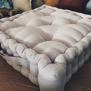 May include: A light gray, square floor cushion with a tufted design. The cushion is made of a soft, textured fabric and has a simple, modern style.