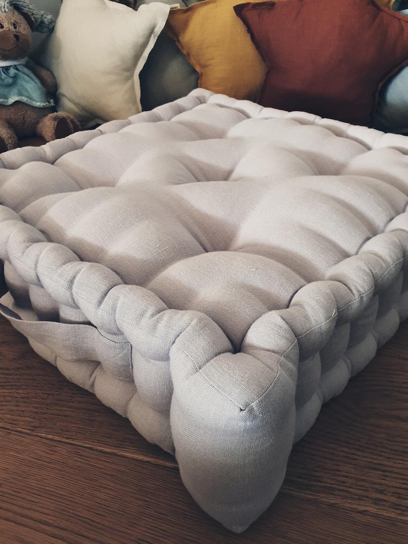 May include: A light gray, square floor cushion with a tufted design. The cushion is made of a soft, textured fabric and has a simple, modern style.