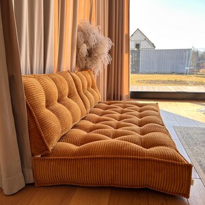 May include: A large, mustard yellow corduroy floor cushion with a tufted design. The cushion is placed in front of a window with beige curtains.