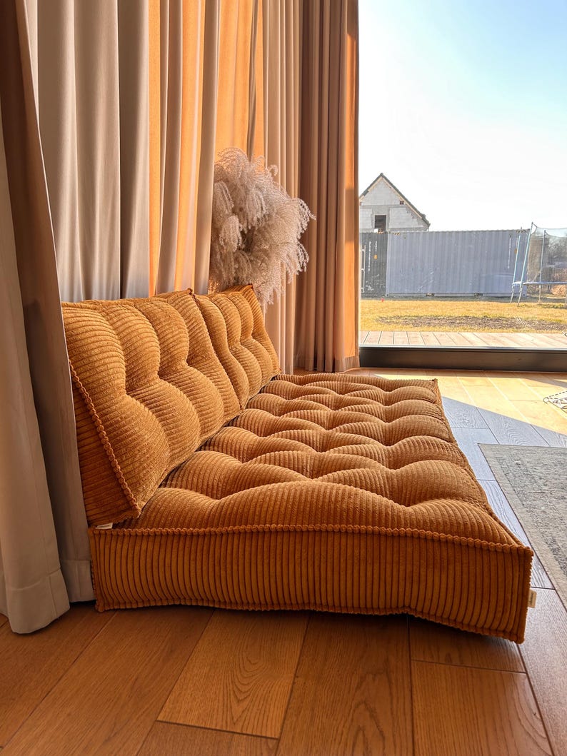 May include: A large, mustard yellow corduroy floor cushion with a tufted design. The cushion is placed in front of a window with beige curtains.