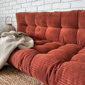 K&ouml;nnte beinhalten: Ein rostfarbenes Cord-Sofa mit getufteter R&uuml;ckenlehne und Sitzfl&auml;che. Eine cremefarbene Strickdecke liegt auf dem Sofa. Das Sofa steht auf einem gewebten Jute-Teppich, mit einer wei&szlig;en Backsteinmauer im Hintergrund.