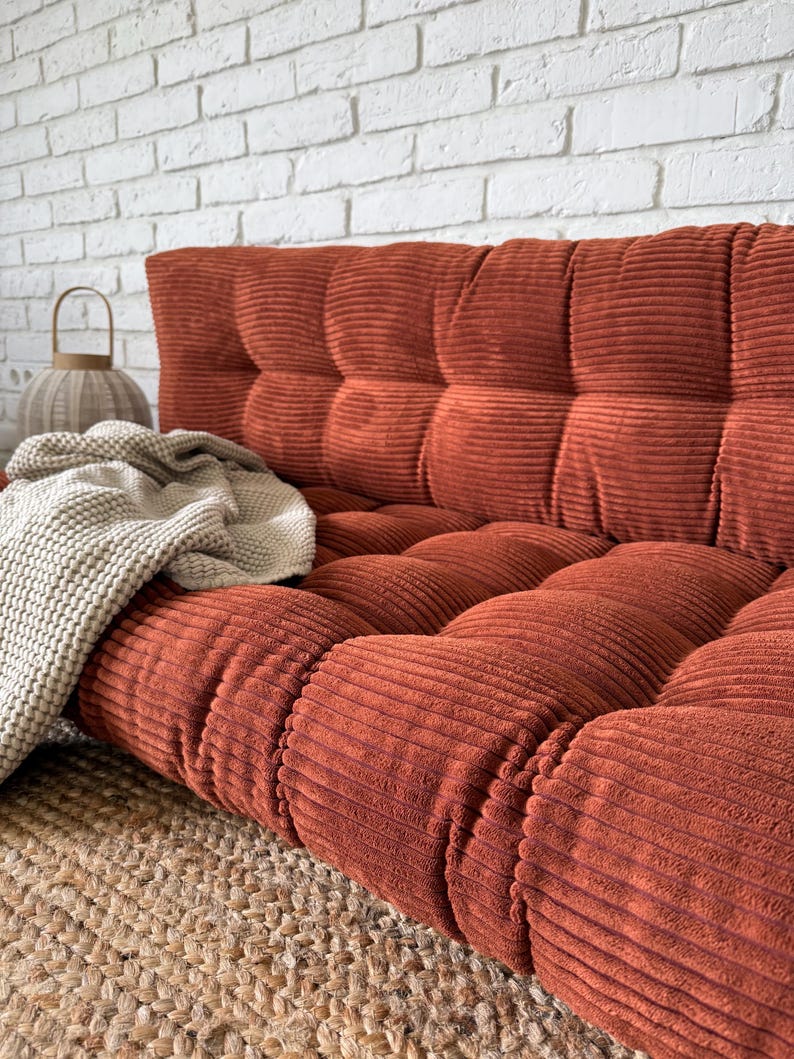 K&ouml;nnte beinhalten: Ein rostfarbenes Cord-Sofa mit getufteter R&uuml;ckenlehne und Sitzfl&auml;che. Eine cremefarbene Strickdecke liegt auf dem Sofa. Das Sofa steht auf einem gewebten Jute-Teppich, mit einer wei&szlig;en Backsteinmauer im Hintergrund.