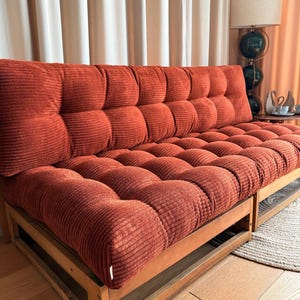 K&ouml;nnte beinhalten: Ein rostfarbenes Cord-Sofa mit Holzrahmen. Das Sofa hat ein getuftetes Design auf den R&uuml;cken- und Sitzkissen. Der Holzrahmen hat ein einfaches, rechteckiges Design. Das Sofa steht vor einem beigen Vorhang.