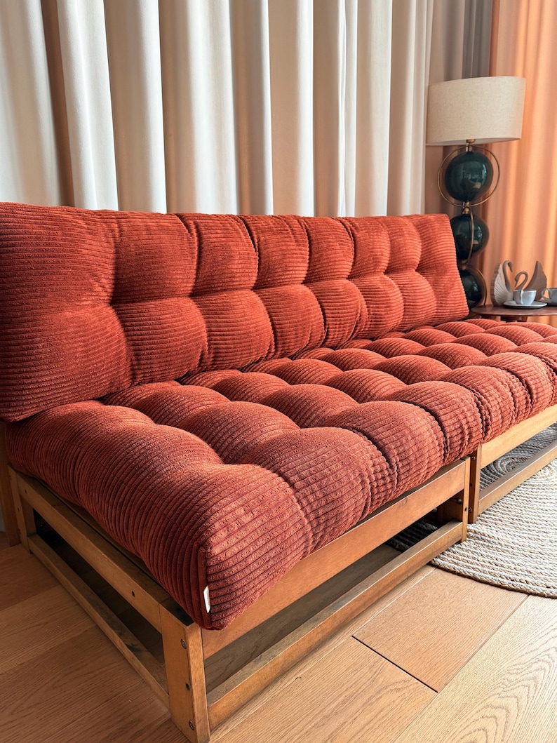 K&ouml;nnte beinhalten: Ein rostfarbenes Cord-Sofa mit Holzrahmen. Das Sofa hat ein getuftetes Design auf den R&uuml;cken- und Sitzkissen. Der Holzrahmen hat ein einfaches, rechteckiges Design. Das Sofa steht vor einem beigen Vorhang.