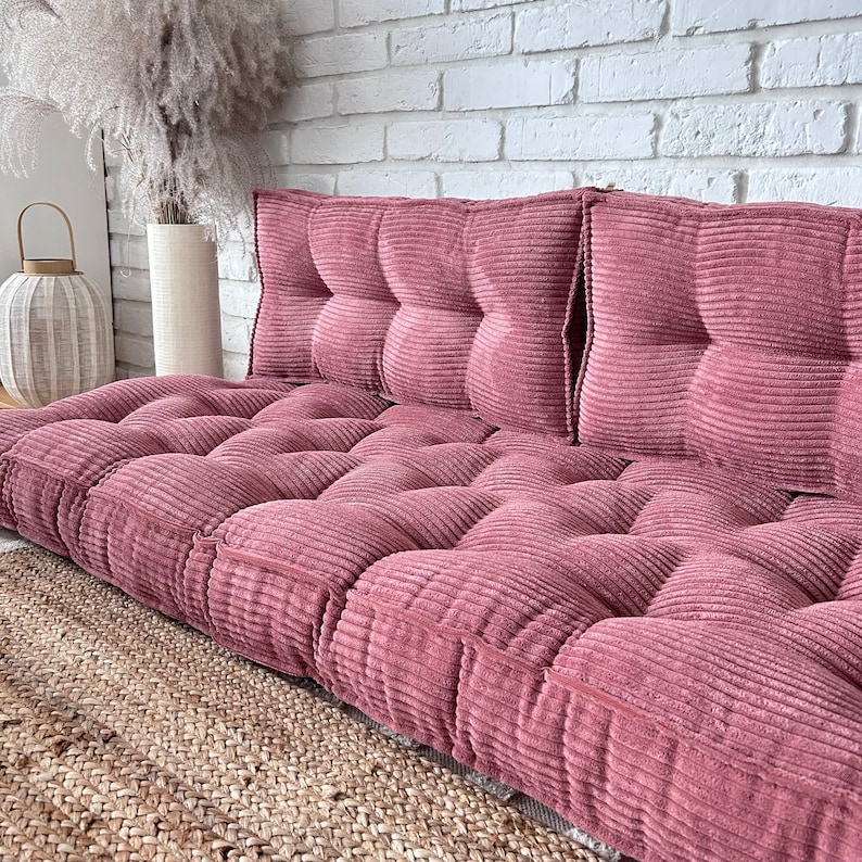 Op de afbeelding: Een roze corduroy vloer kussen met een gestoffeerd ontwerp. Het kussen bestaat uit meerdere vierkanten en is perfect om elke kamer een vleugje comfort en stijl te geven.
