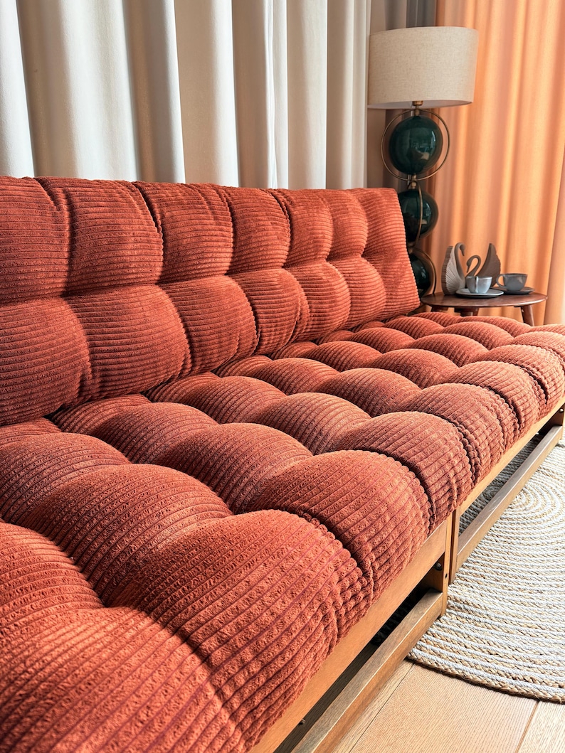 Może przedstawiać: Sofa z sztruksu w kolorze rdzy z pikowanym wzorem. Sofa ma drewnianą ramę i znajduje się w pokoju z neutralnymi zasłonami i lampą. Poduszki sofy są ułożone we wzór kratki.