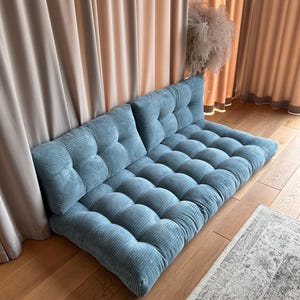 May include: A blue corduroy floor cushion with tufted buttons. The cushion is made up of two separate pieces that can be used as a sofa or a bed.