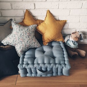 May include: A blue floor cushion with a tufted design and a handle. The cushion is surrounded by pillows, including two yellow star-shaped pillows and a floral patterned pillow.