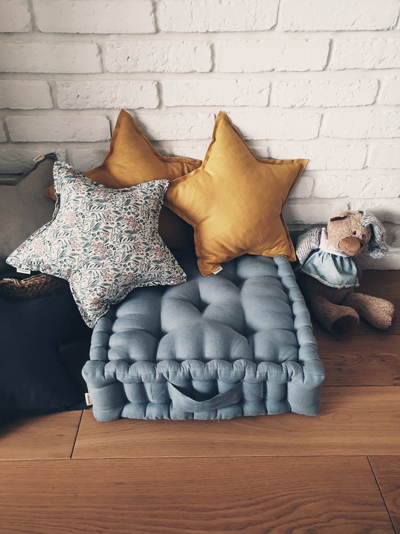 May include: A blue floor cushion with a tufted design and a handle. The cushion is surrounded by pillows, including two yellow star-shaped pillows and a floral patterned pillow.