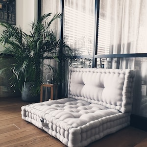 Op de afbeelding: Een groot, lichtgrijs, gestoffeerd vloerkleed met een ritssluiting. Het kussen is gemaakt van een zachte, linnenachtige stof en is perfect om te loungen of te mediteren. Het kussen ligt op een houten vloer voor een raam met witte gordijnen.