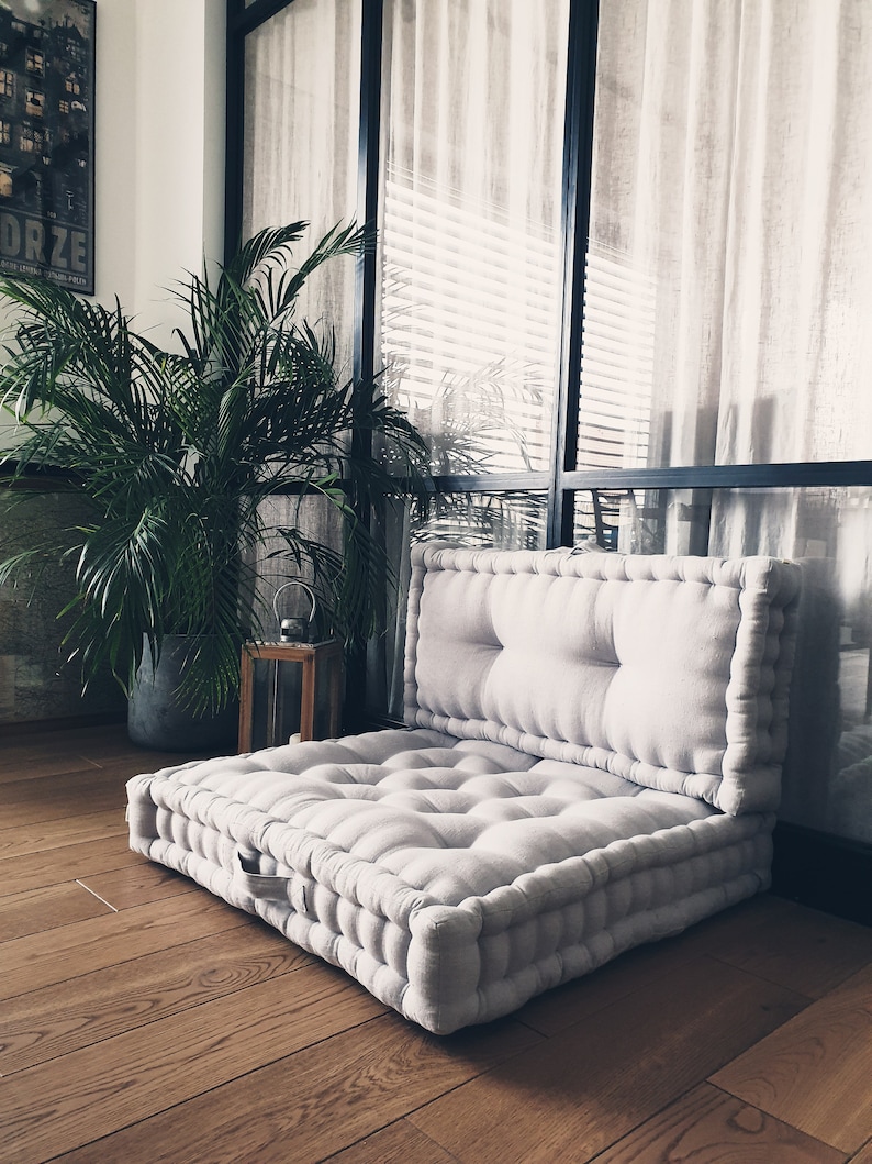 Op de afbeelding: Een groot, lichtgrijs, gestoffeerd vloerkleed met een ritssluiting. Het kussen is gemaakt van een zachte, linnenachtige stof en is perfect om te loungen of te mediteren. Het kussen ligt op een houten vloer voor een raam met witte gordijnen.
