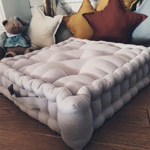 May include: A light gray floor cushion with tufted detailing. The cushion is made of linen and has a square shape. It is sitting on a wooden floor with several decorative pillows behind it.