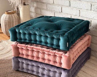 May include: Three square floor cushions stacked on top of each other. The top cushion is teal velvet, the middle cushion is dusty rose velvet, and the bottom cushion is a dark gray velvet. The cushions are all tufted and have a white piping around the edges.