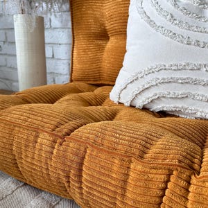Op de afbeelding: Een mosterdgele corduroy vloercushion met een wit kussen met franjes. Het kussen is gestoffeerd en heeft een knoopdetail.