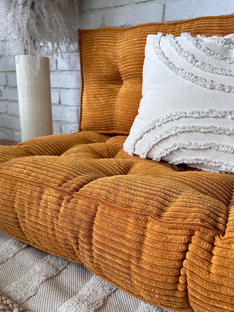Op de afbeelding: Een mosterdgele corduroy vloercushion met een wit kussen met franjes. Het kussen is gestoffeerd en heeft een knoopdetail.