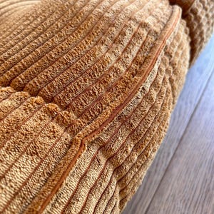 May include: Close-up of a brown corduroy cushion with a seam running down the center. The cushion is made of a soft, textured fabric.