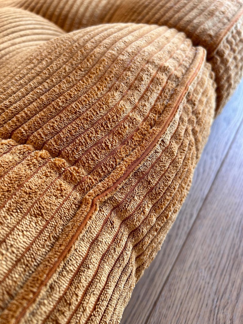 May include: Close-up of a brown corduroy cushion with a seam running down the center. The cushion is made of a soft, textured fabric.