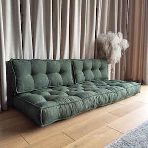 Könnte beinhalten: Ein grünes, getuftetes Bodensofa mit niedrigem Profil. Das Sofa hat eine rechteckige Form mit einer Reihe von quadratischen, getufteten Kissen. Der Stoff hat ein strukturiertes Aussehen. Das Sofa steht auf einem Holzboden.