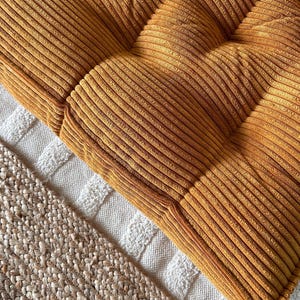 Op de afbeelding: Een mosterdgele corduroy vloercushion met een gestoffeerd ontwerp. Het kussen ligt op een wit en beige kleed.