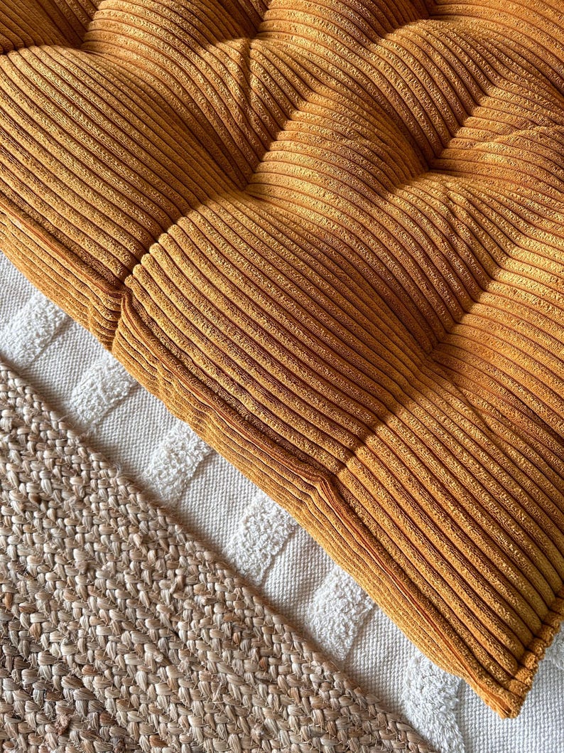 Op de afbeelding: Een mosterdgele corduroy vloercushion met een gestoffeerd ontwerp. Het kussen ligt op een wit en beige kleed.