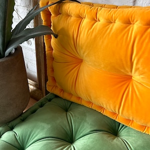May include: A bright yellow velvet cushion with button tufting sits on top of a green velvet cushion with button tufting. Both cushions have a soft, plush texture.